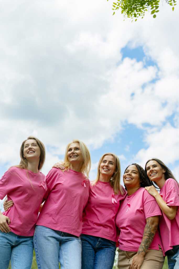 Women smiling and supporting breast cancer awareness, wearing pink ribbons and shirts outdoors