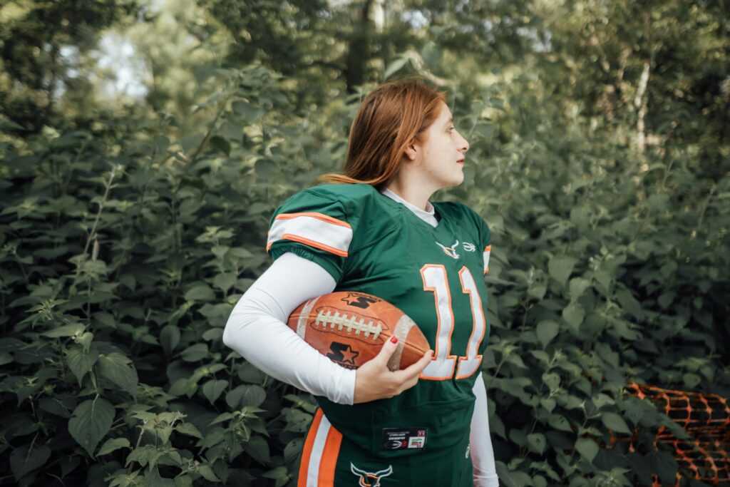 A woman in football gear stands confidently outdoors holding a football.
